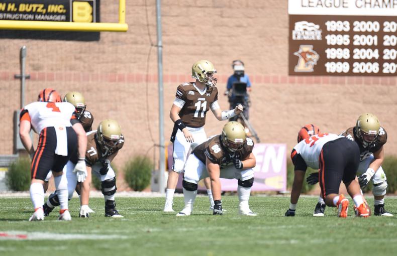 Three linebackers dressed in brown uniforms on the line, prepared for the hand off with the quarterback calling directions behind them.