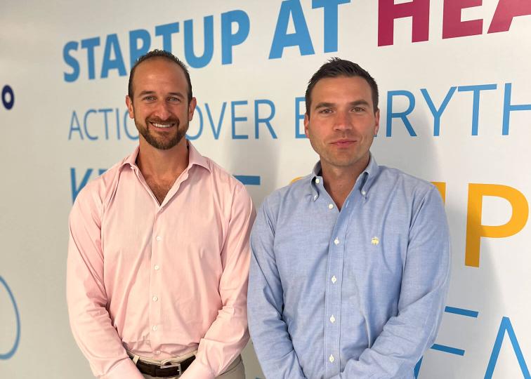 Business partners wearing button down shirts stand smiling in front of a wall emblazoned with brightly colored words.
