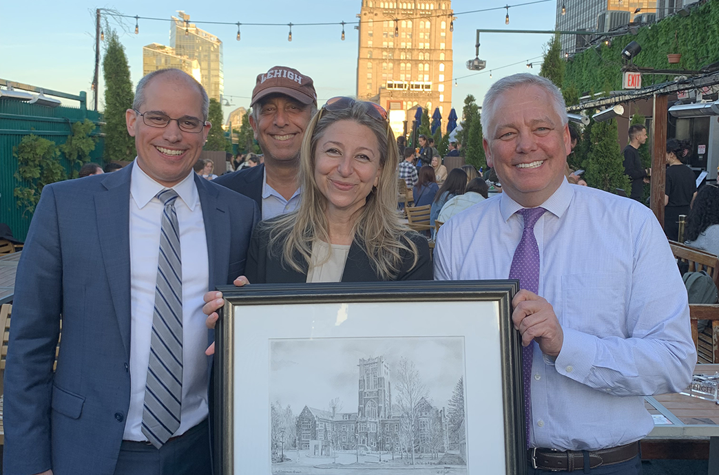 a group of alumni holding up a framed drawing of the University Center, and smiling at an outdoor event.