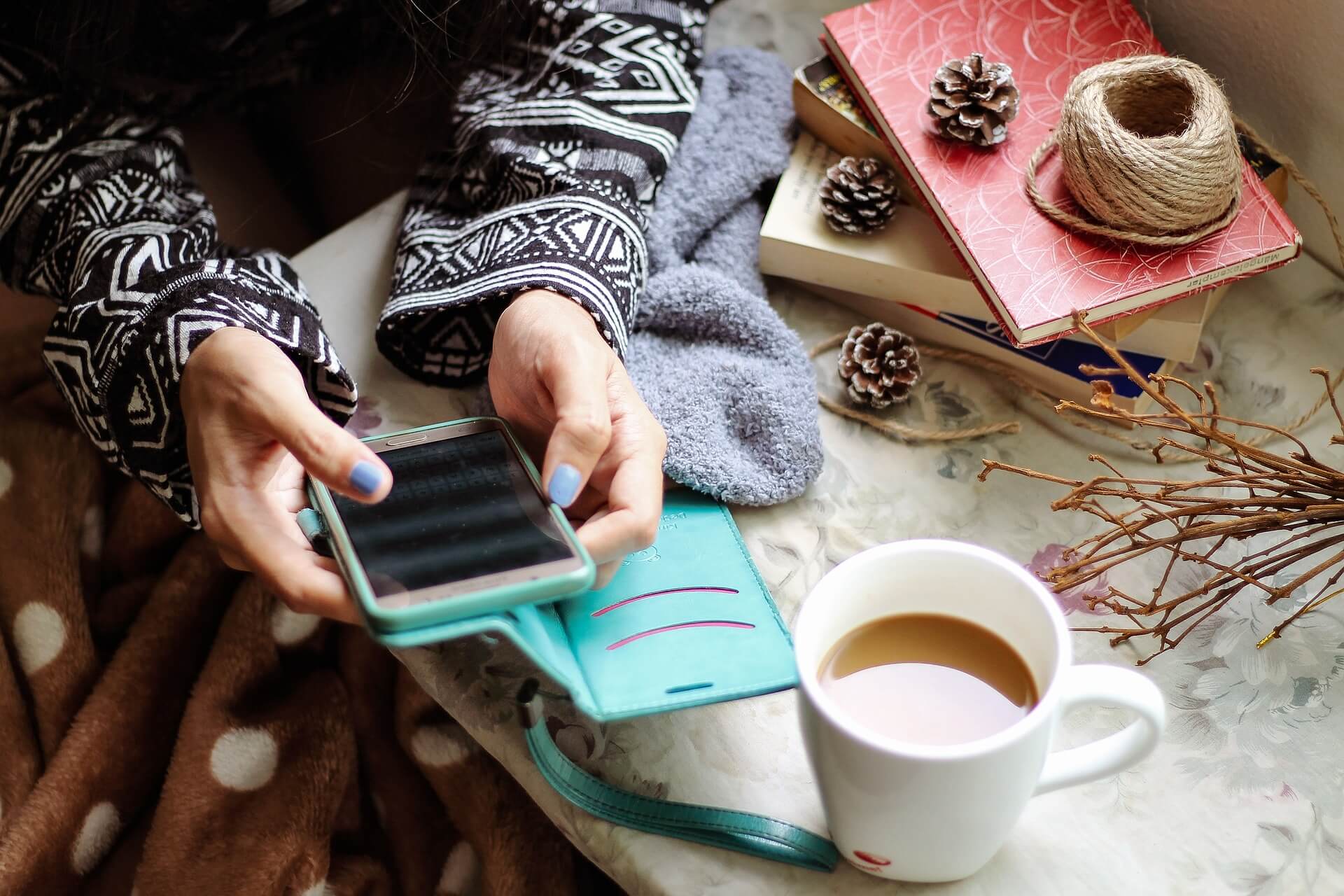 A close-up of a woman's hands typing on her smart phone with a white mug of coffee