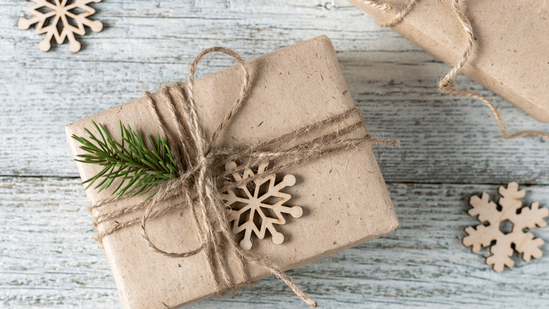 High angle view of brown paper wrapped gift, tied with natural twine, a sprig of a pine branch, and small wooden snowflake ornament.