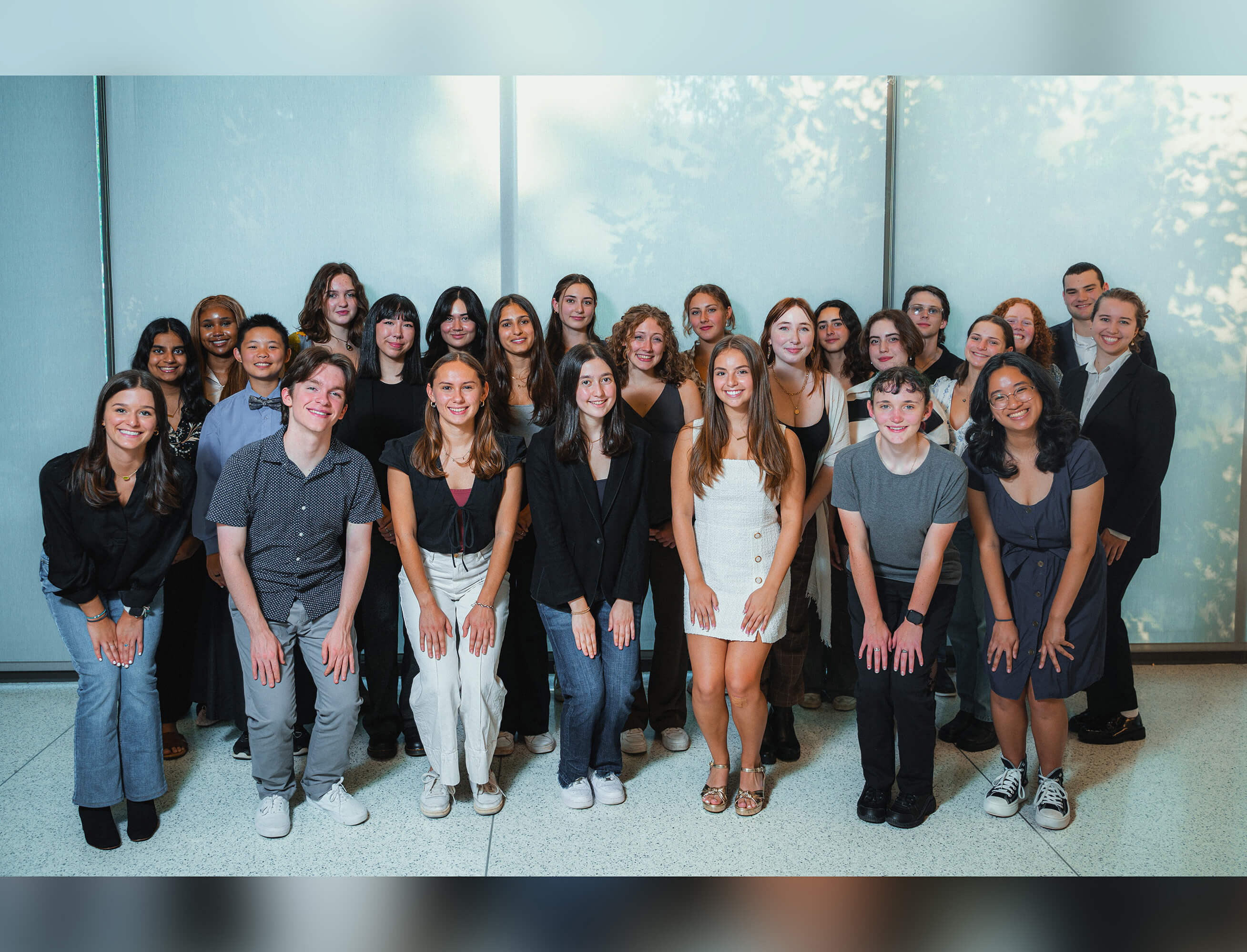 Standing in 3 rows with the front row squatting with hands on their knees, Students smile while posing against a wall of windows. 