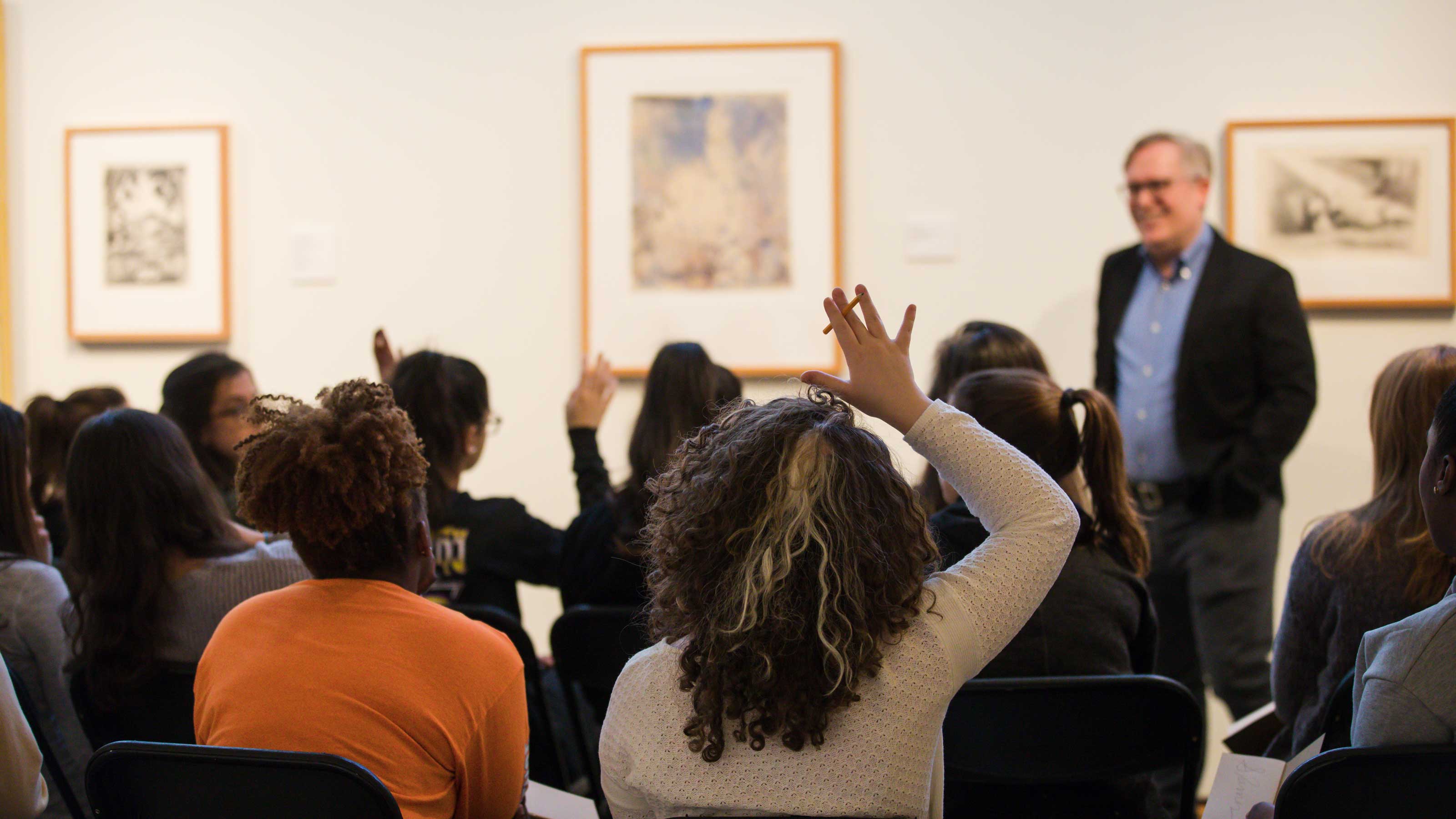 Seated audience with raised hands viewed from behind, viewing art and a speaker at the front of the room.