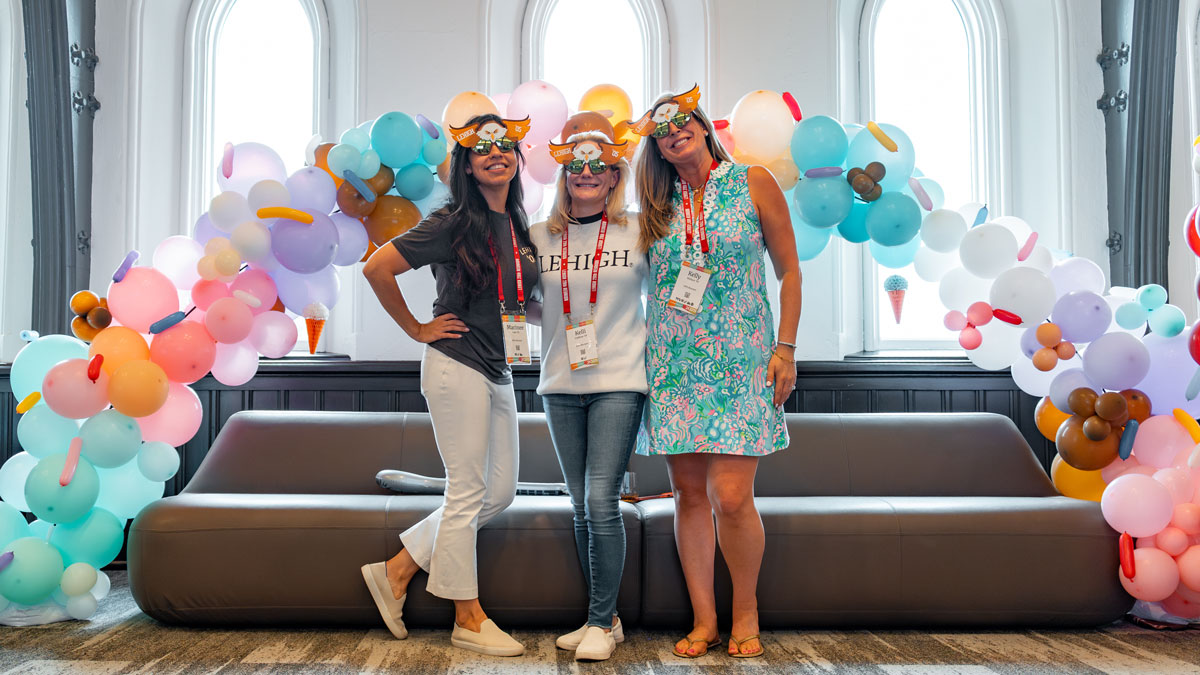 Three women wearing fun masks pose together in front of a balloon arch with pastel-colored balloons and a grey couch.