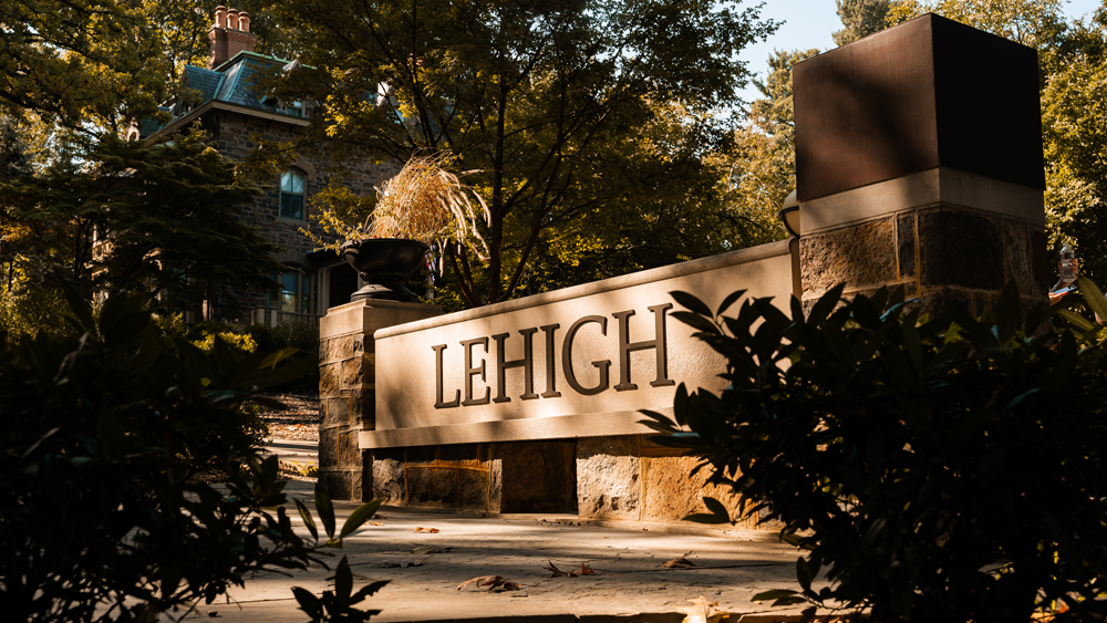 Stone bench at Lehigh University with "Lehigh" engraved on it, surrounded by lush greenery and historic campus architecture in the background, bathed in warm sunlight.