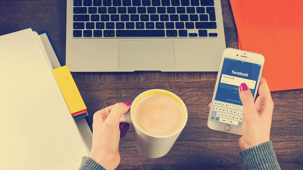 A pair of hands holding a mug of coffee and a smartphone opened to Facebook with a laptop keyboard and files in view.