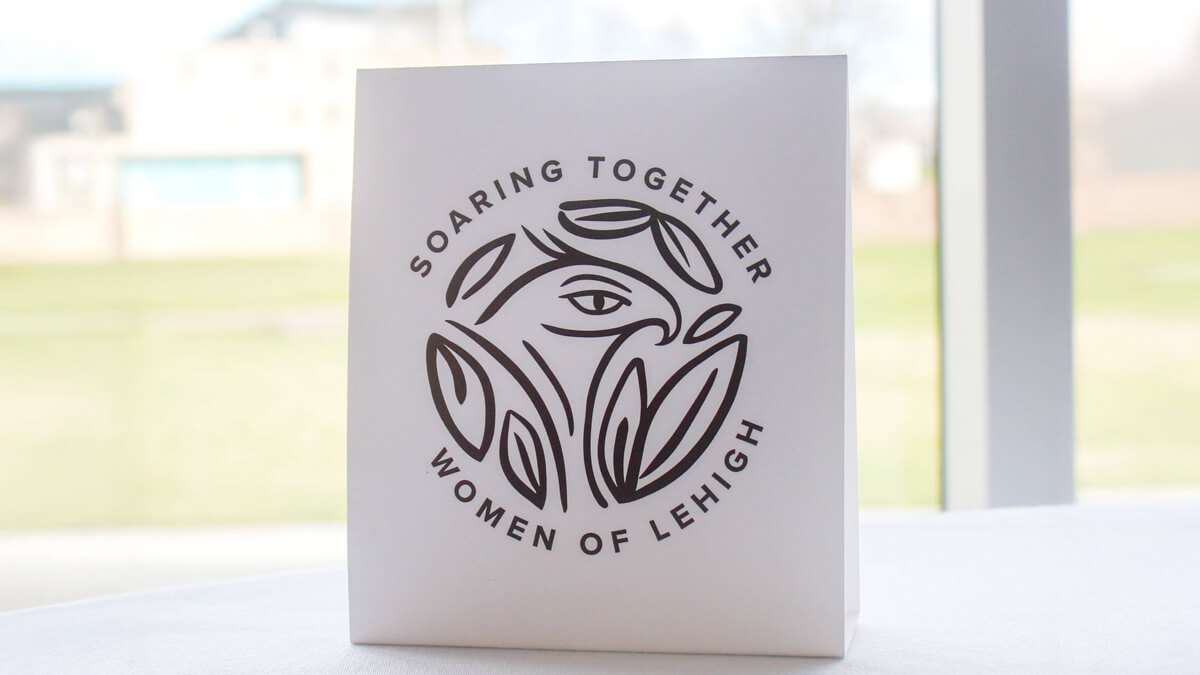 A white card displays a stylized design with the text “SOARING TOGETHER WOMEN OF LEHIGH” arranged in a circle around the graphic. The card sits on a white surface with a blurred outdoor background.