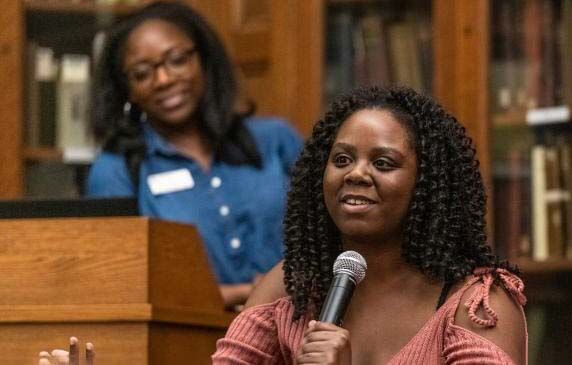 A woman speaks into a microphone while another woman smiles in the background.