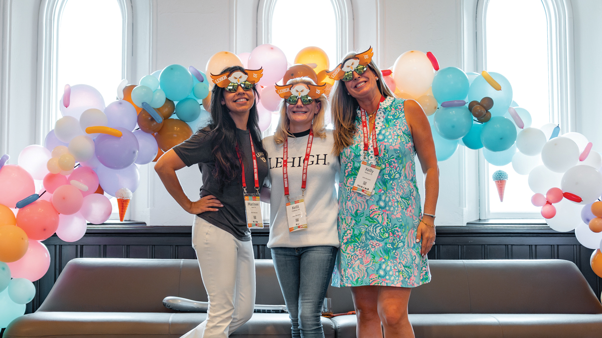 Three women wearing fun masks pose together in front of a balloon arch with pastel-colored balloons and a grey couch.