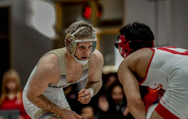 Two male wrestlers face each other on a mat, wearing singlets and headgear. One athlete wears beige and gold for Lehigh, the other wears red and white for Cornell.