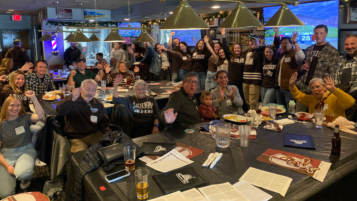 A group of alumni cheer in a restaurant while televisions behind them show the Rivalry football game.