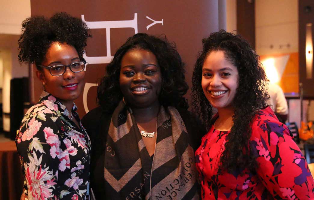 Three women stand before a standing floor banner and smile.