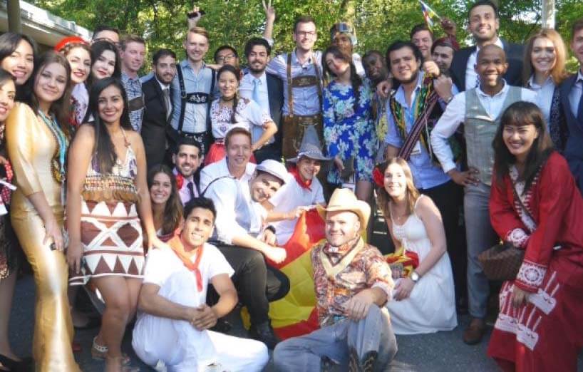 A group of college-aged people pose together outdoors, many of them smiling and wearing traditional or cultural clothing from various countries.