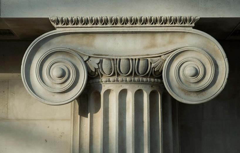 A close-up of a white stone Ionic column capital, showing its characteristic volutes (scrolls) on either side of a fluted column shaft.