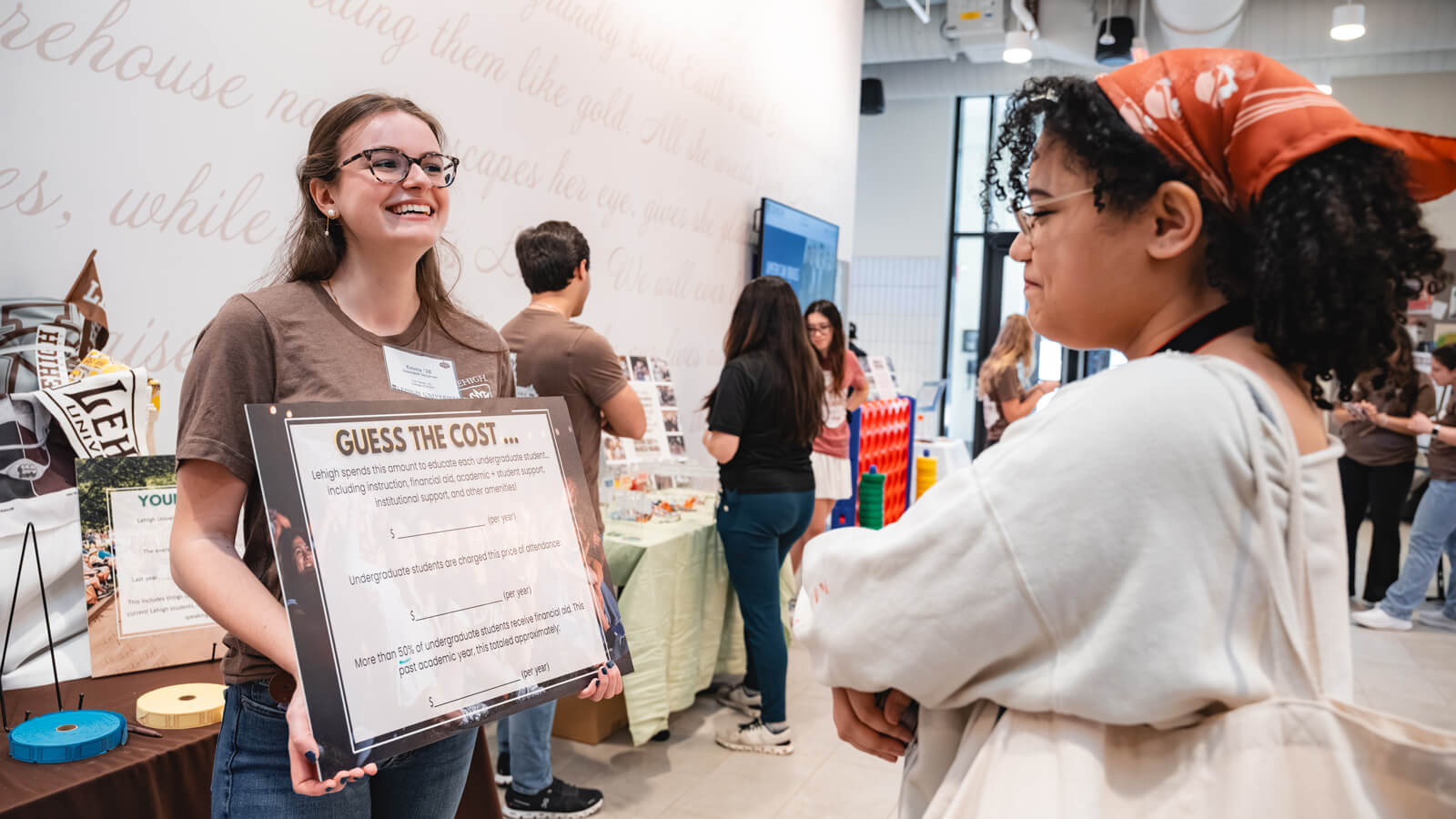 A smiling student at Lehigh University holds a "Guess the Cost" sign while talking to another student during a Giving Day event. The sign includes prompts about education costs, tuition, and financial aid.