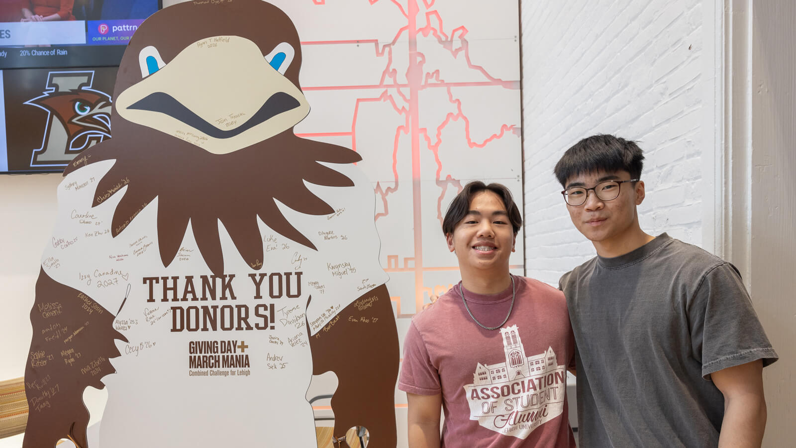 Two male Lehigh students stand next to a large Lehigh mountain hawk mascot cutout at a Lehigh University "Giving Day" event.