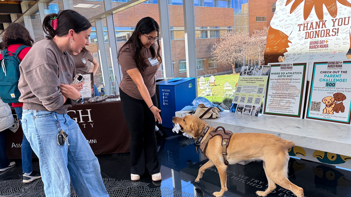 Two Lehigh University students smile and reach out to pet a yellow Labrador wearing a harness. They are standing in a sunlit campus building next to a "Giving Day + March Mania" table featuring "Thank You Donors" signs and information about fundraising.