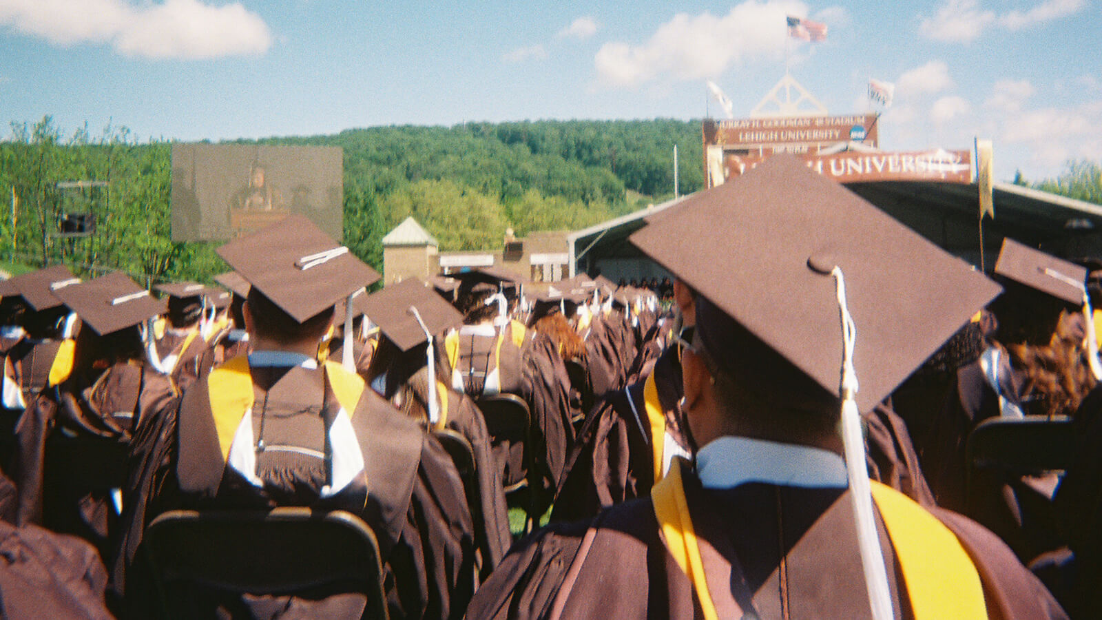 Graduates in caps and gowns face a stage at Lehigh University, with an outdoor screen showing the ceremony and flags visible.