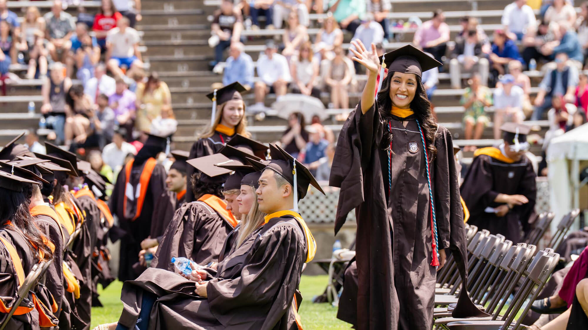 A female graduate in a brown cap and gown waves and smiles while walking past rows of seated students during a commencement ceremony.