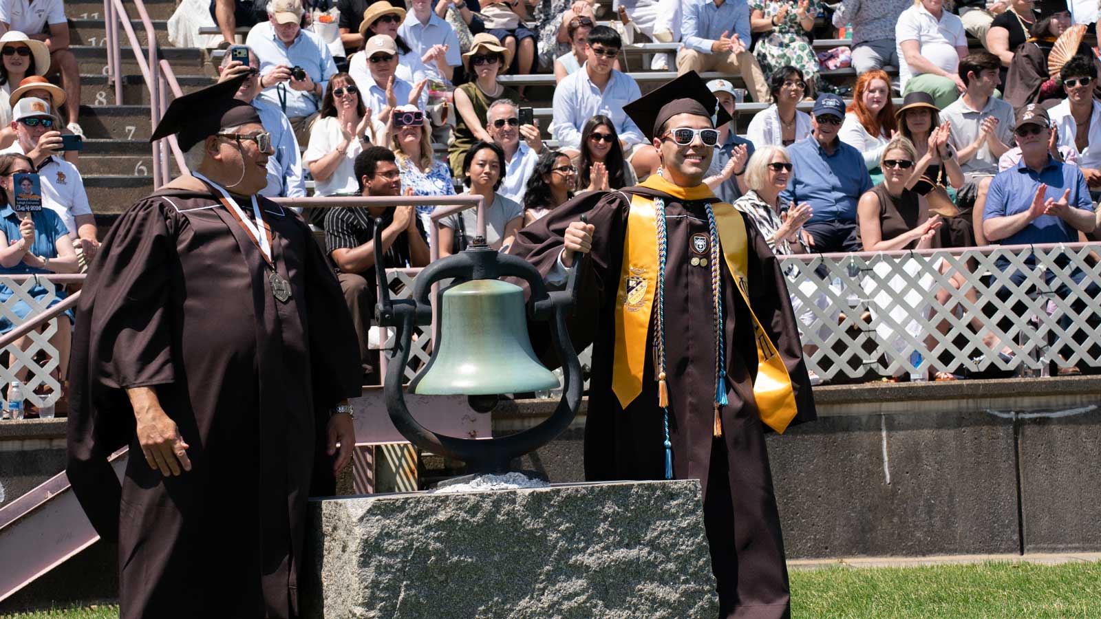 A graduate in brown and gold regalia rings a large bronze bell during a commencement ceremony with a crowd watching.