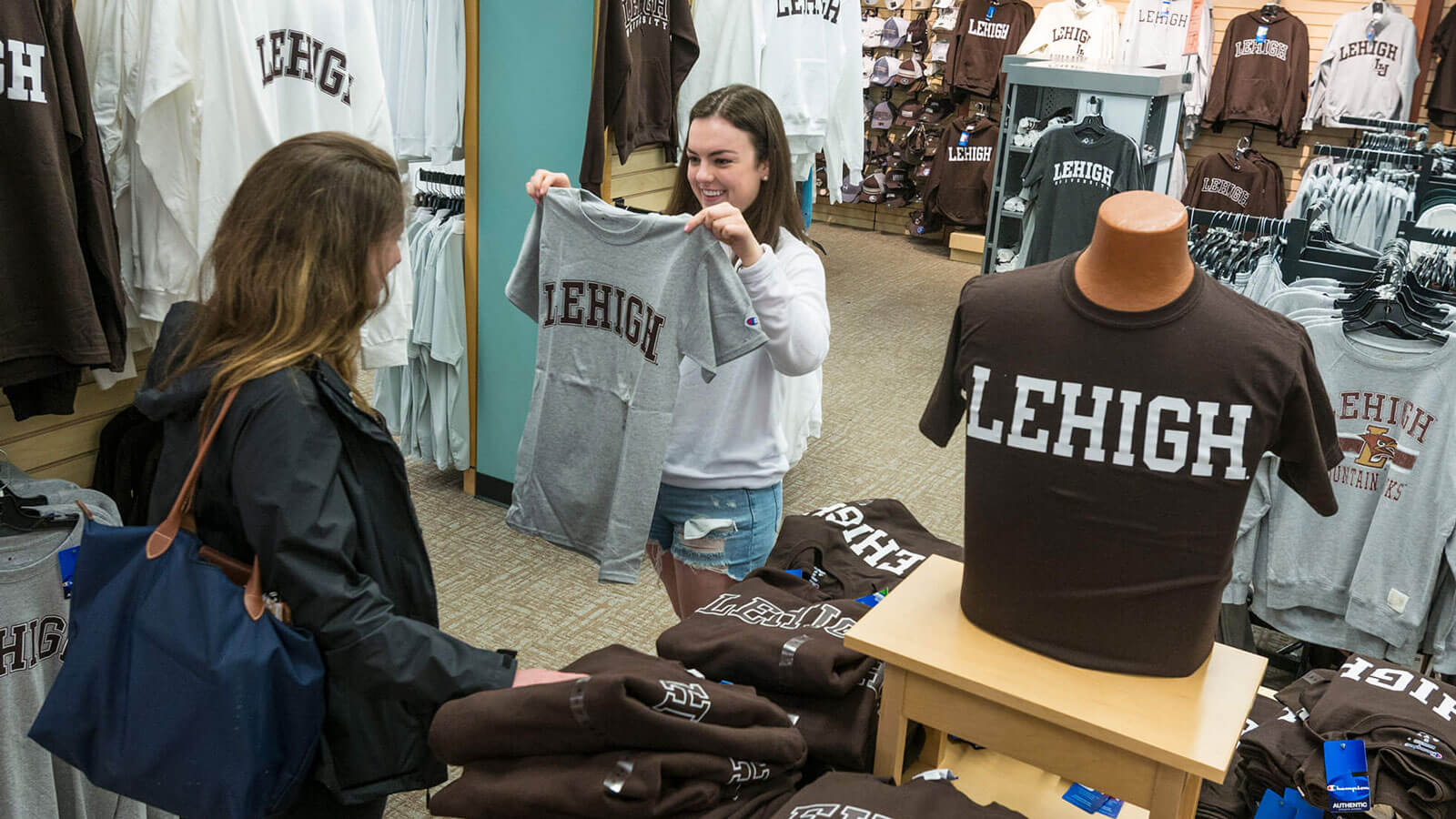 Daughter holds up a gray shirt that says "Lehigh" in brown lettering while mom admires it amid racks of Lehigh clothing.