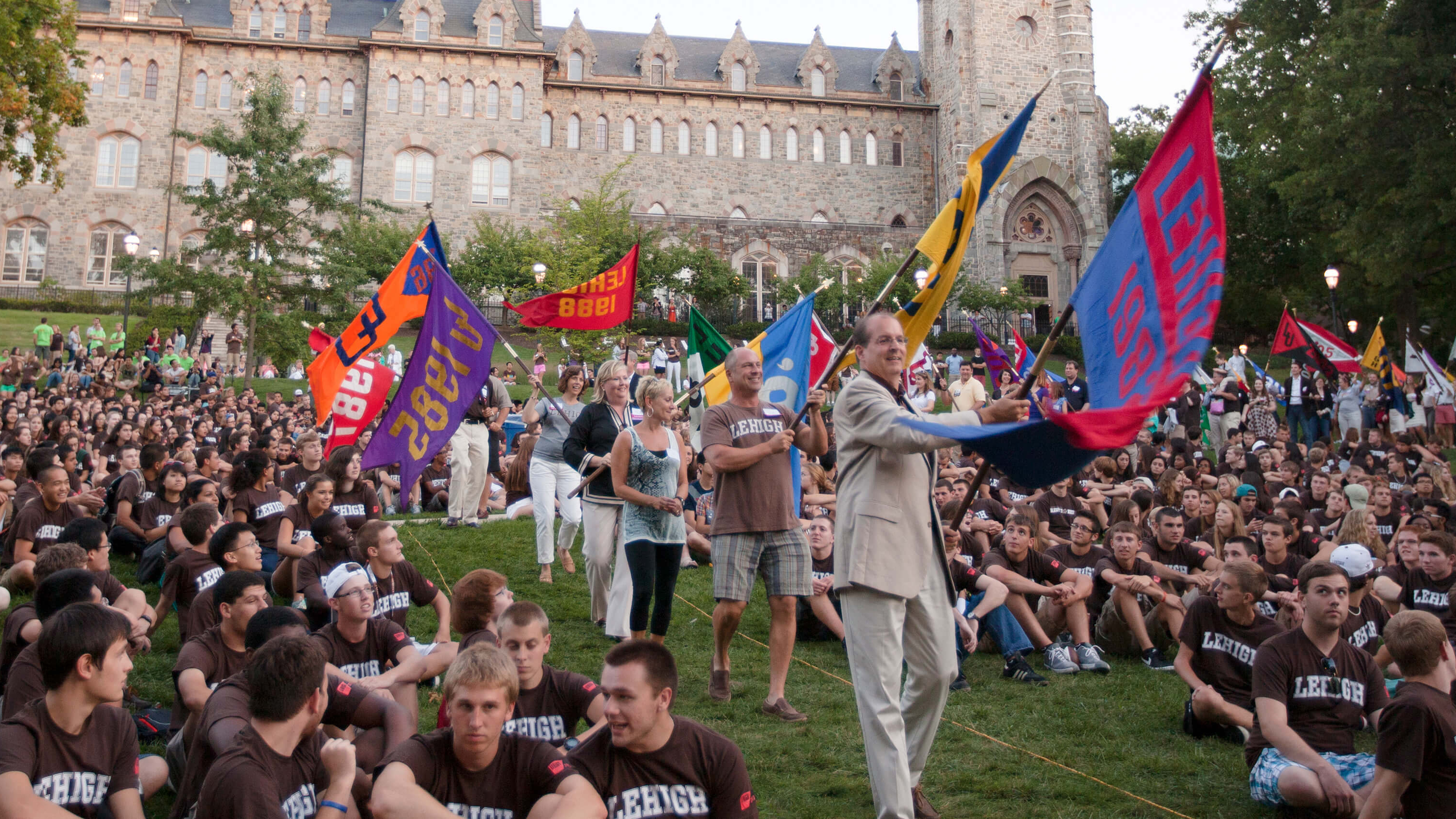 Alumni from the ’80s carry class flags and parade between crowds of students seated on the grass on the front lawn of the University Center and who are all wearing brown Lehigh tshirts.