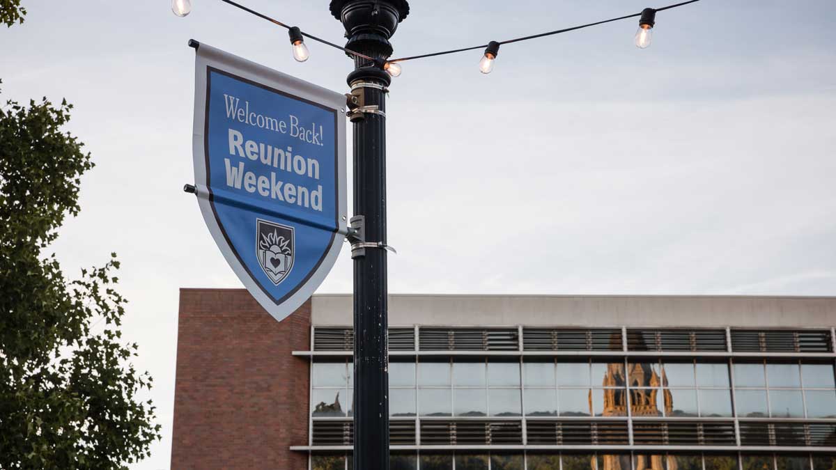 A blue shield-shaped banner hanging from a black lamppost reads "Welcome Back! Reunion Weekend."