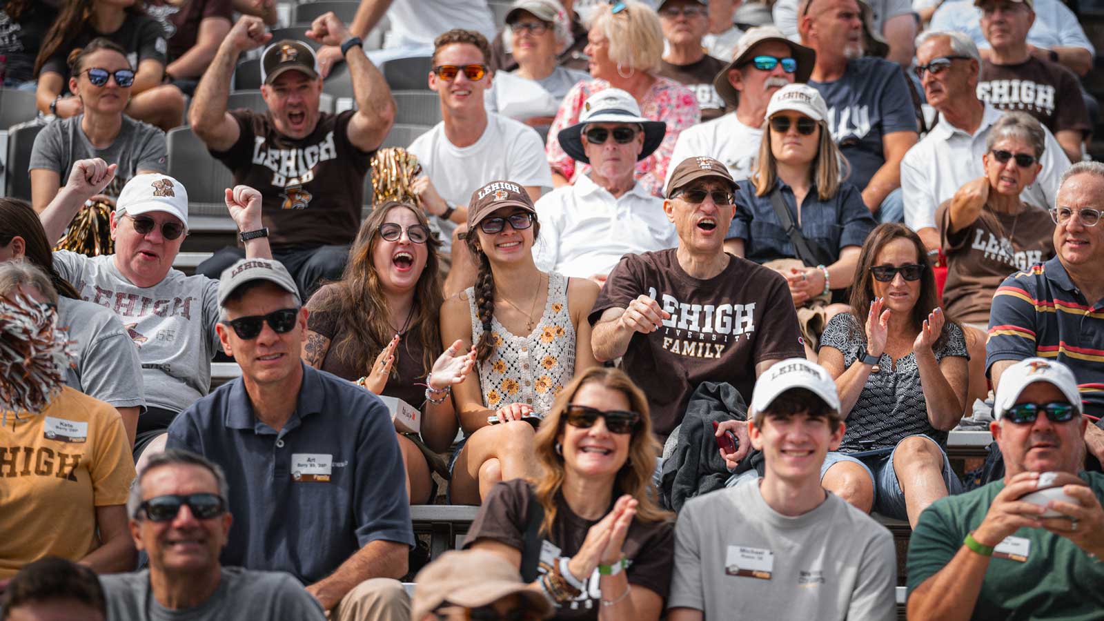 A crowded stadium bleacher filled with enthusiastic fans wearing brown and white Lehigh University gear cheering during a football game.
