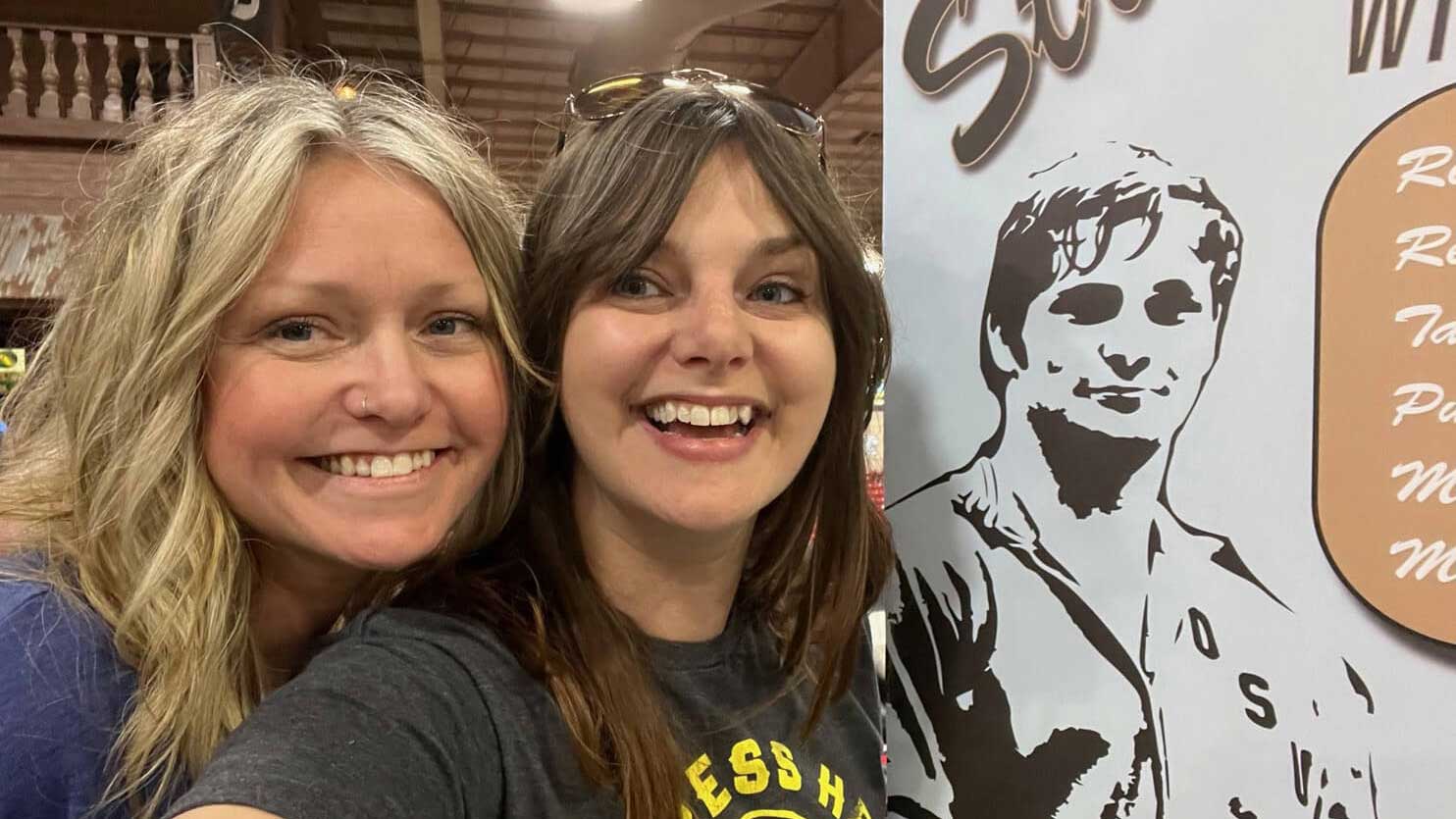 The Strobel sisters pose smiling for a selfie beside a banner with an illustration of their father.
