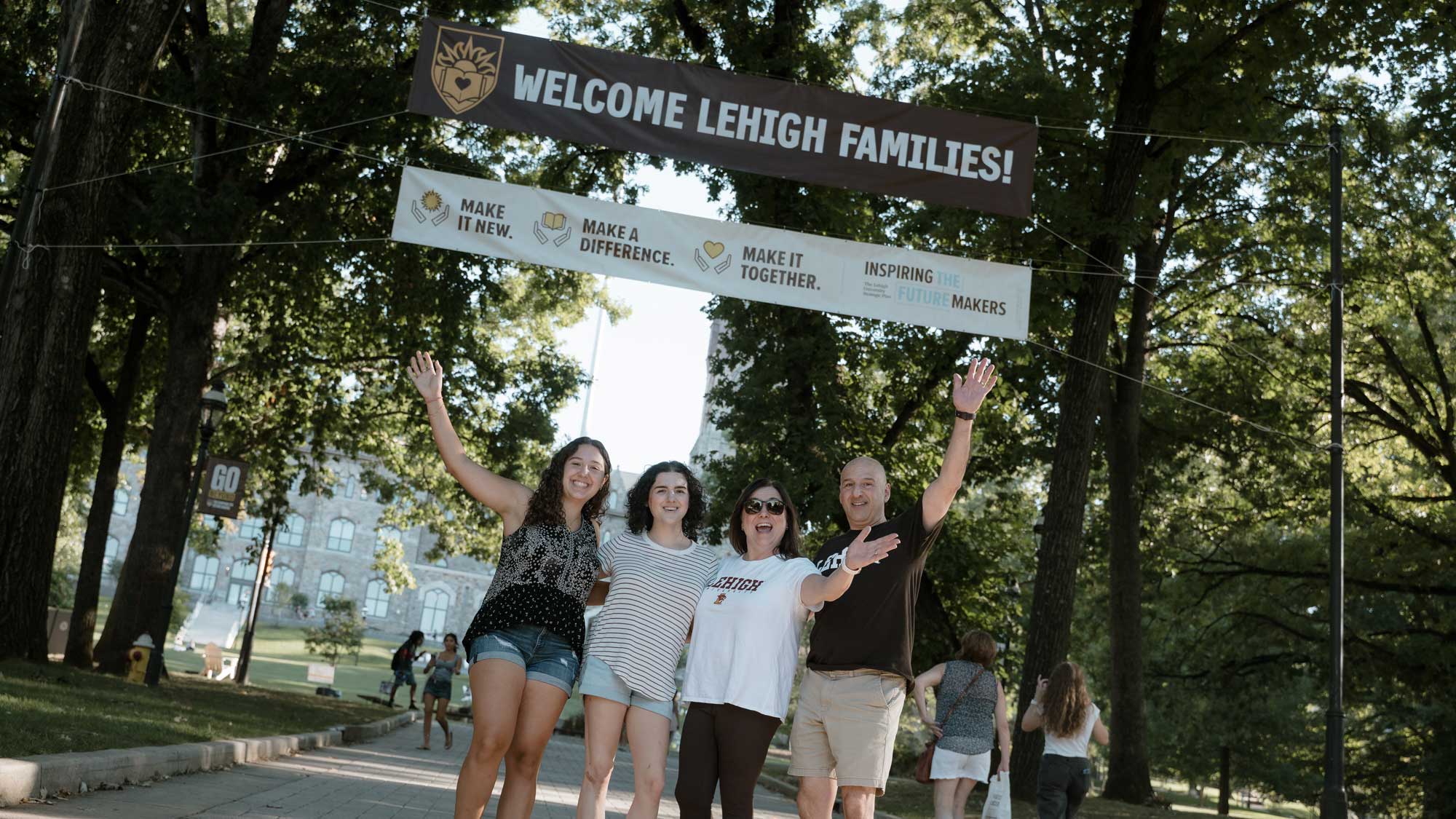 A family of four waves cheerfully beneath a "WELCOME LEHIGH FAMILIES!" banner strung between trees on a campus walkway, with stone campus buildings visible in the background.