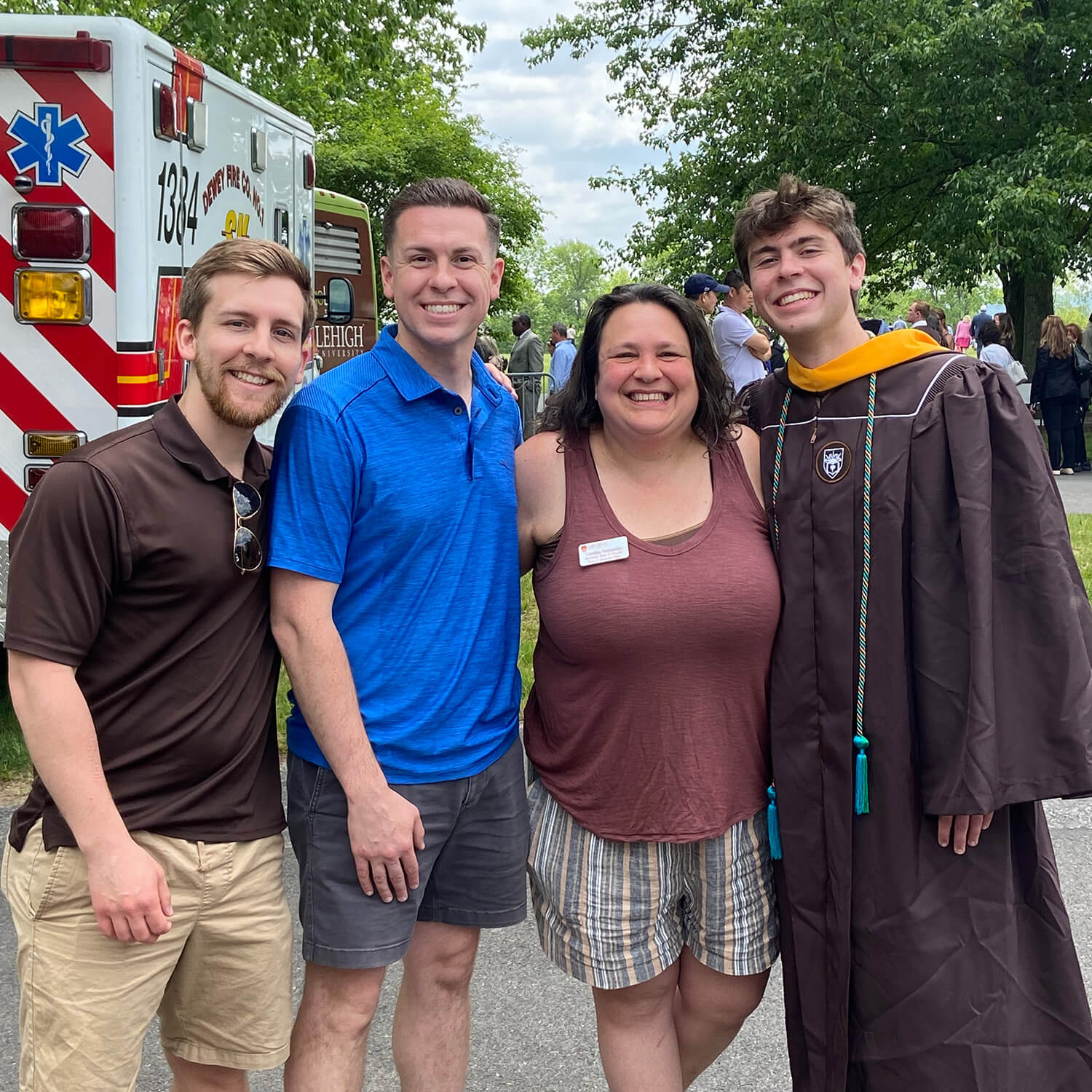 A woman in a brown tank top and shorts stands smiling with a graduate wearing a brown robe and tassels and his two brothers.