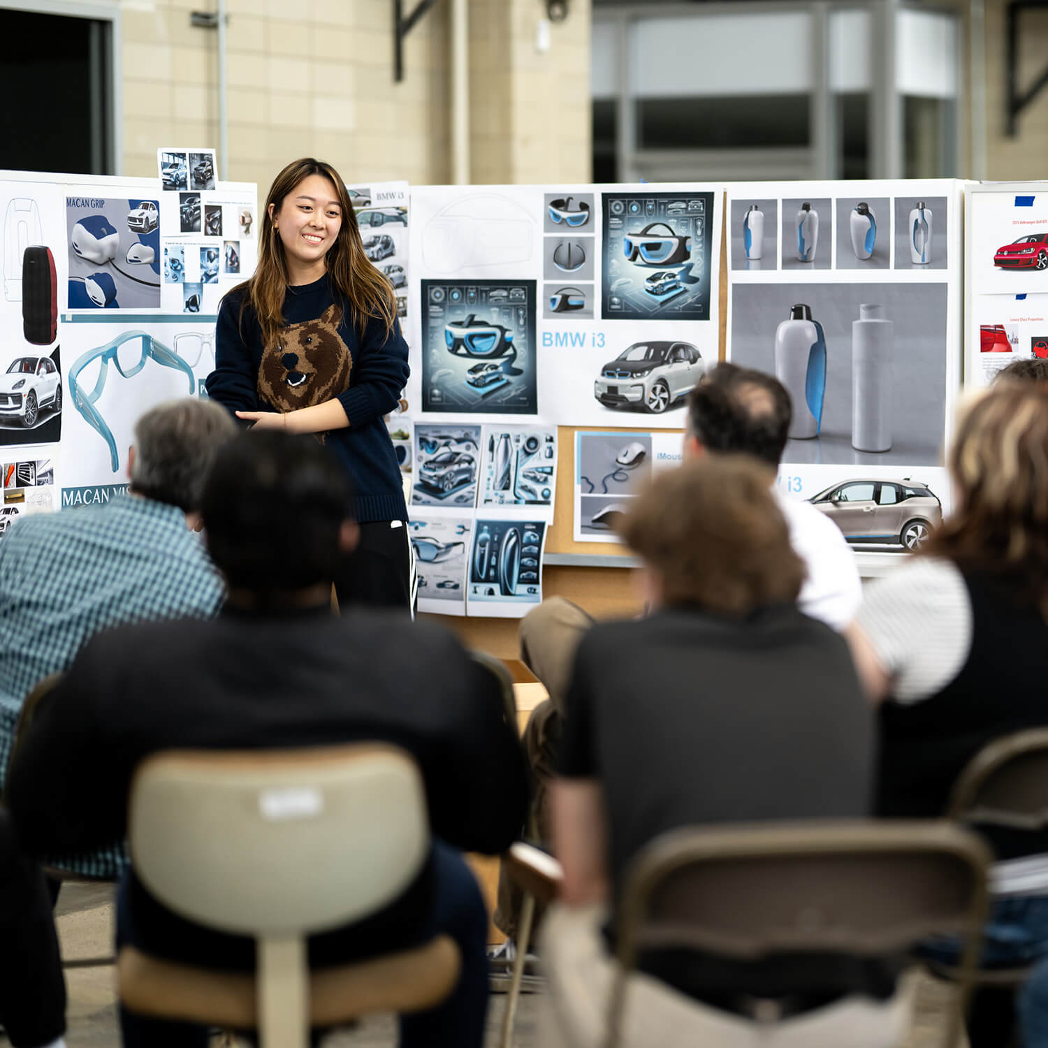 A student presents design work to an audience, standing in front of display boards with product and automotive concepts.