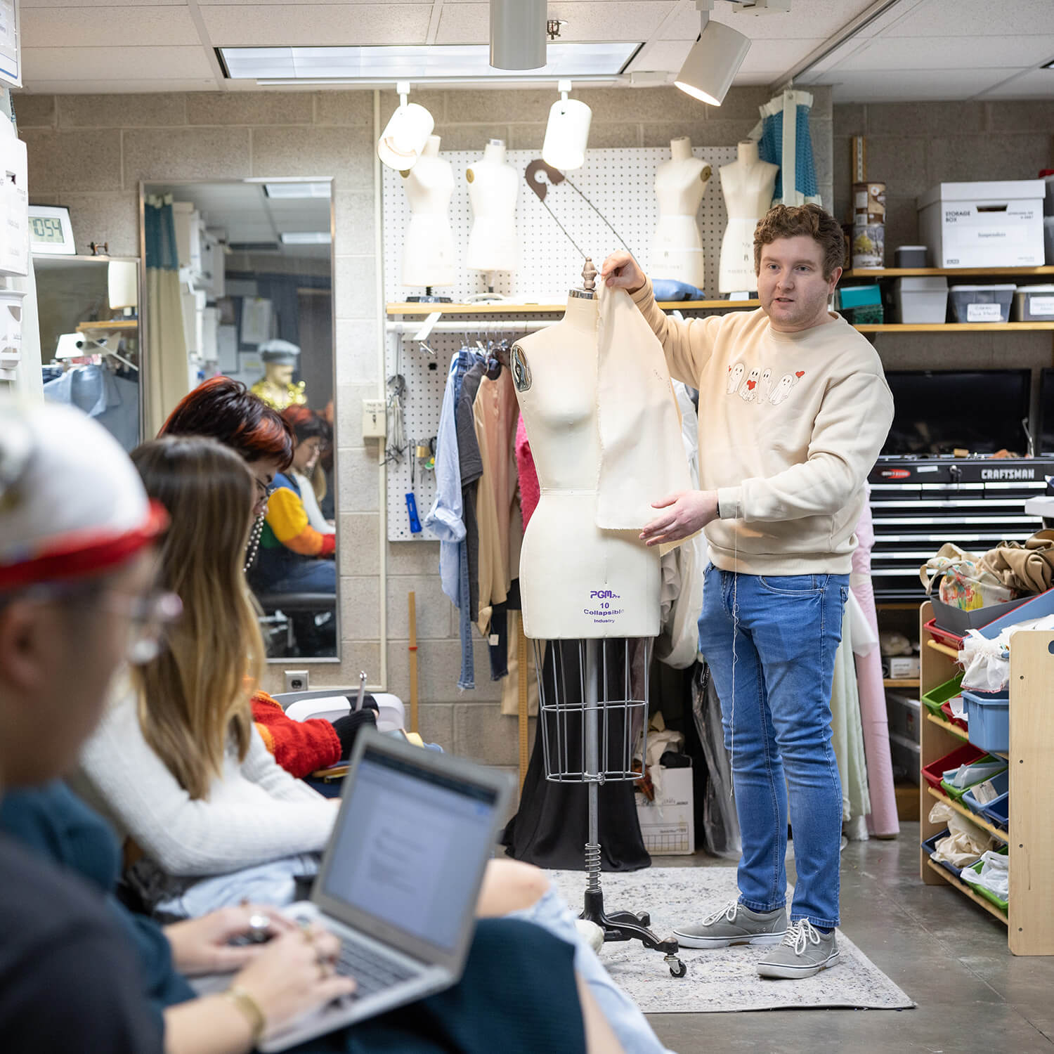 nstructor demonstrates garment construction on a dress form while students observe in a studio.