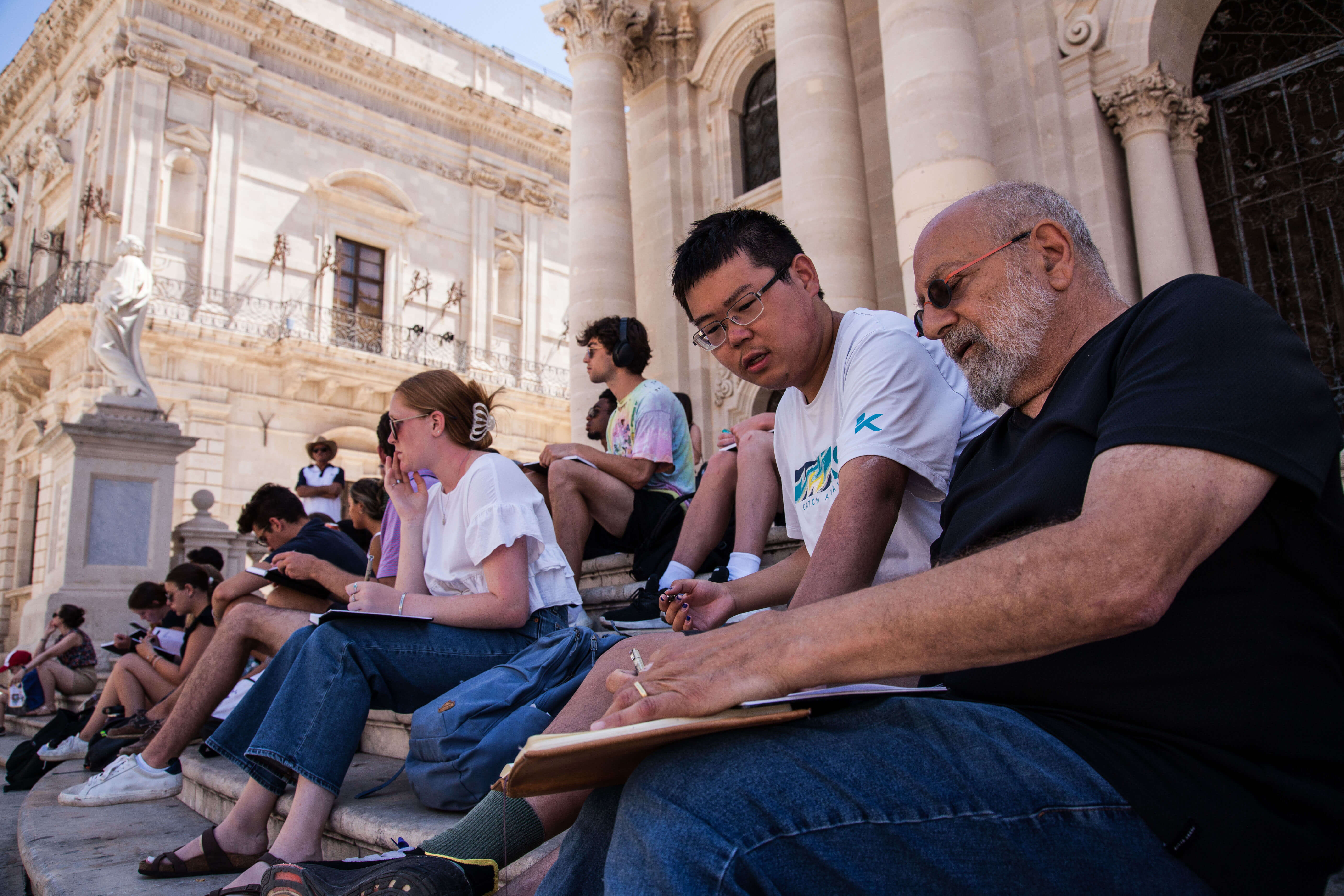 A group of people with notebooks sketch buildings on the steps of a stone building