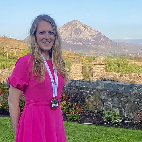 Elle wears a pink dress and a medal around her neck from a recent award. A mountain landscape is behind her