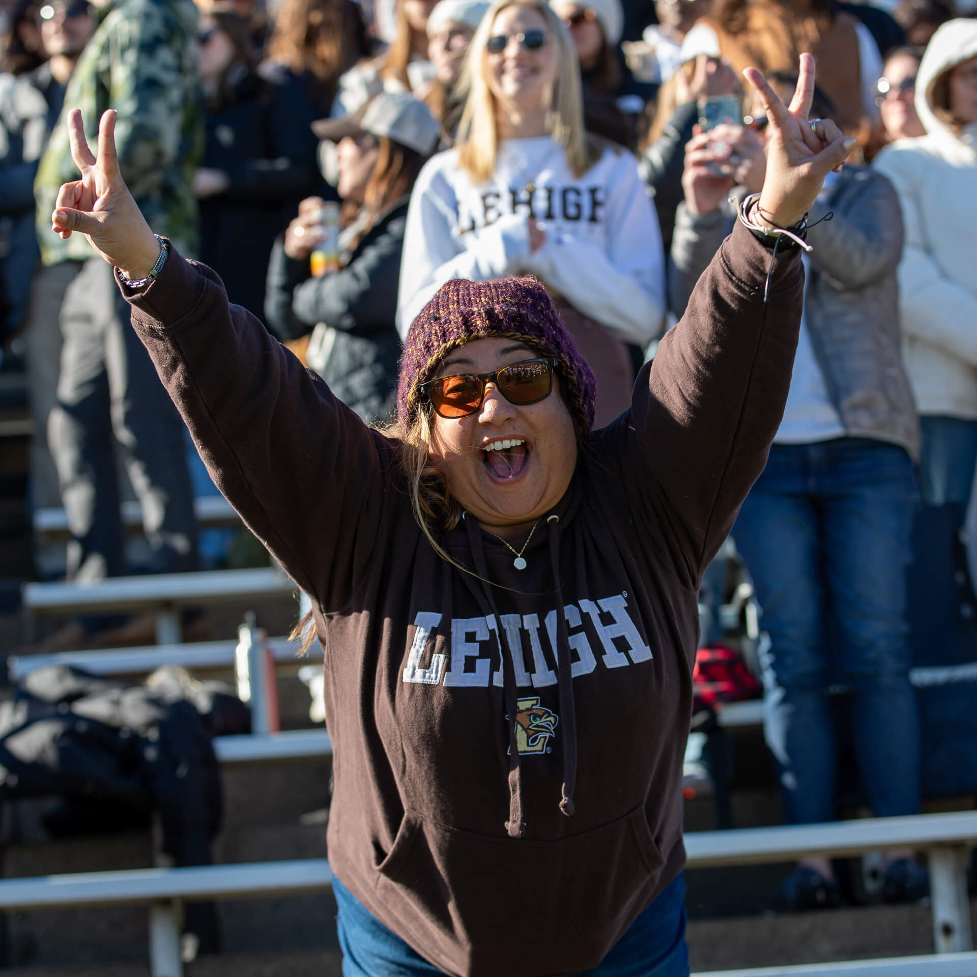 Alumna dressed in brown cheers at a football game, reaches her arms in the air, and makes peace signs with both hands