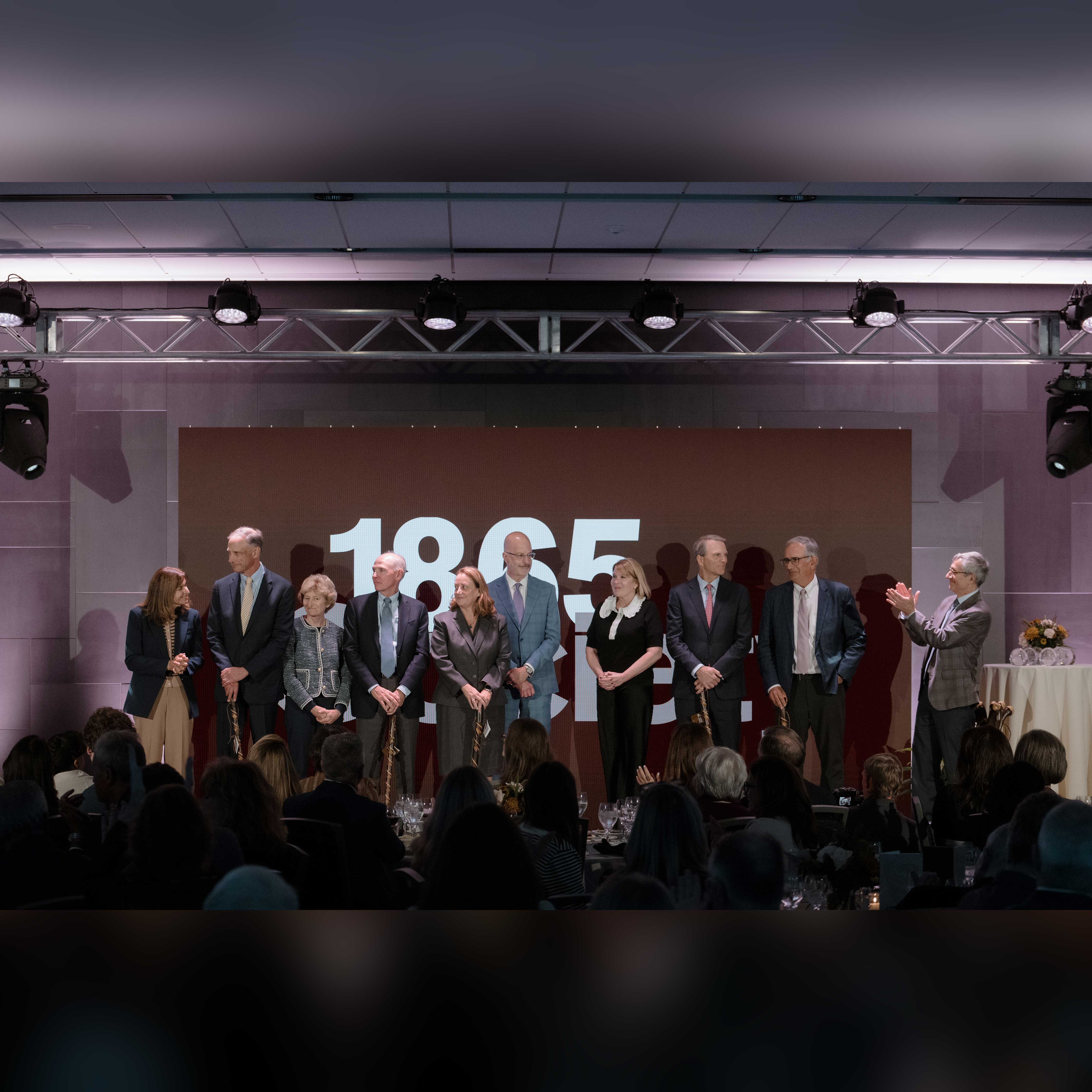 The 1865 Society inductees stand on the stage and hold their honorary canes. President Helble stands to their right and claps.