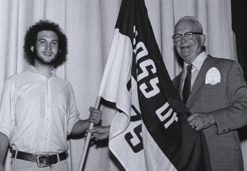 In a black and white photo, a man with dark hair and beard holds a “Class of 1979” flag alongside an older gentleman wearing a suit jacket and glasses.