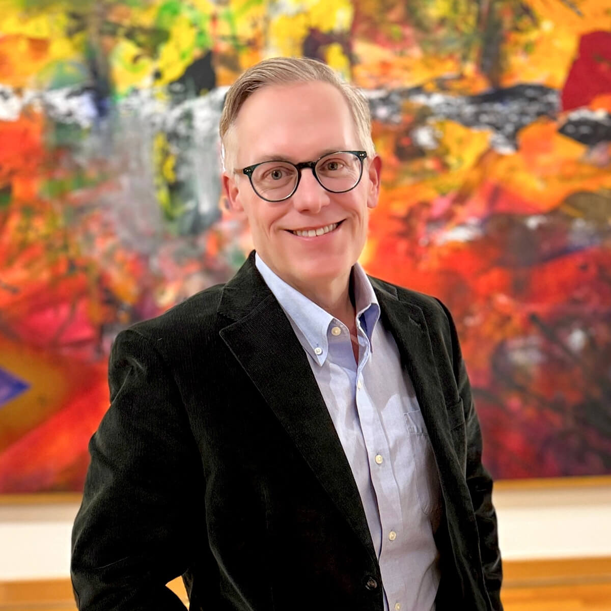 Standing in front of a large, colorful abstract painting, William Crow smiles wearing glasses, a black suit jacket, and button-down collared shirt.