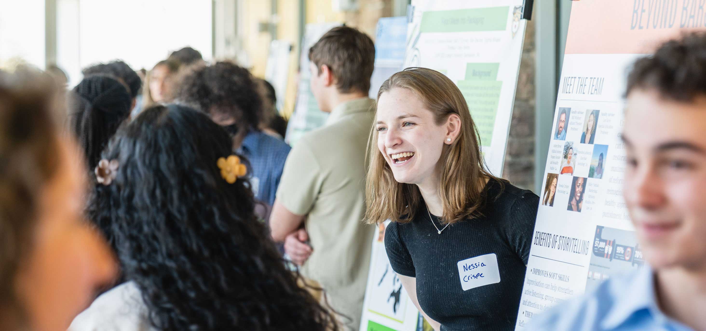 A student smiling in a crowded hallway while presenting her project.