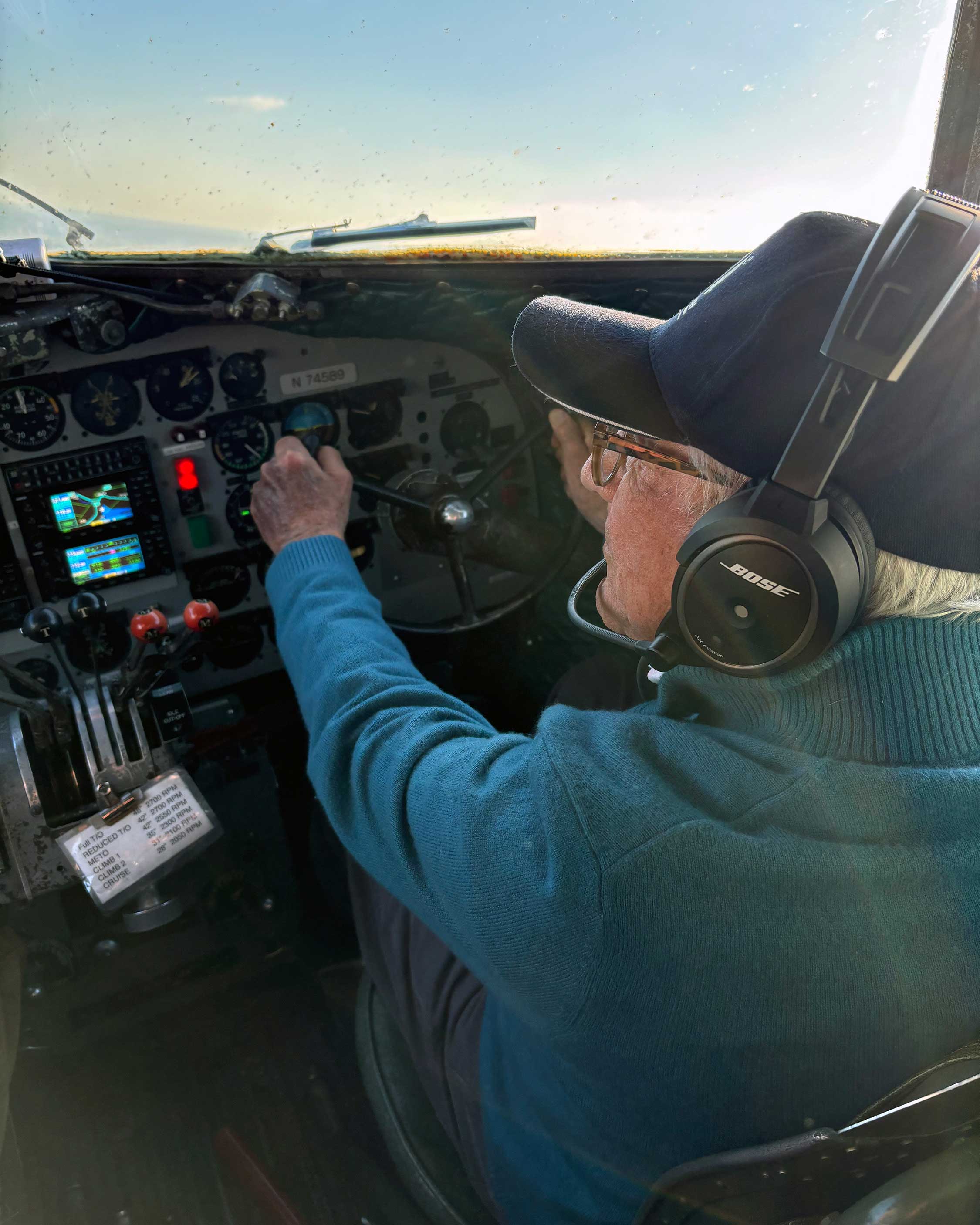 Alan sits in the cockpit of a plane and flies it. He wears a dark blue sweater, black hat, and Bose headphones. The sky is clear.