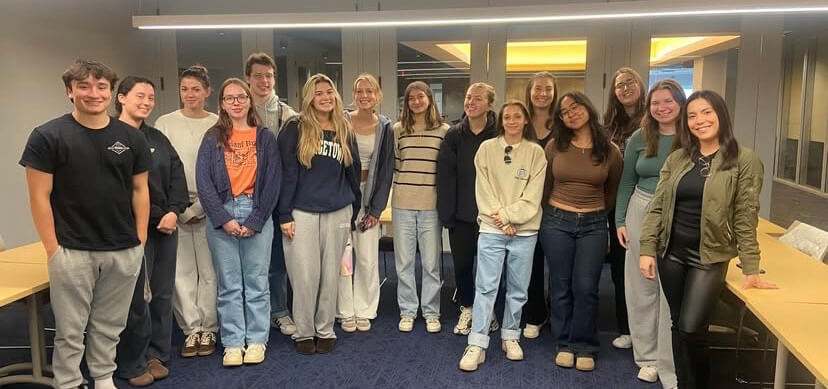  A group of students casually dressed stand in a conference room for a photo, smiling with with Carissa Iacona standing to the far right.