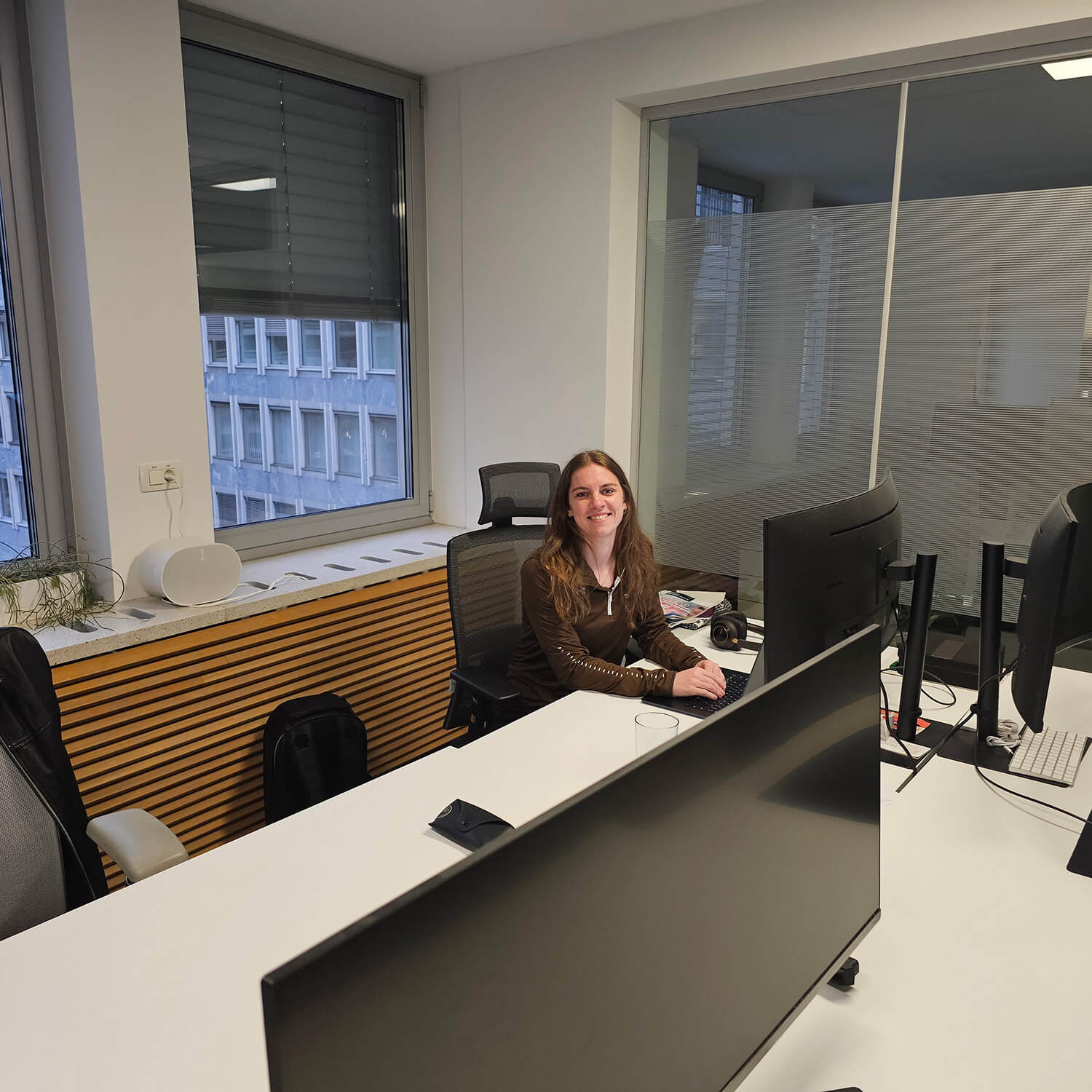 Kaja Skerlj sits smiling in a black office chair at a large desk with computer monitors along the top with a high rise shown in the window behind her.