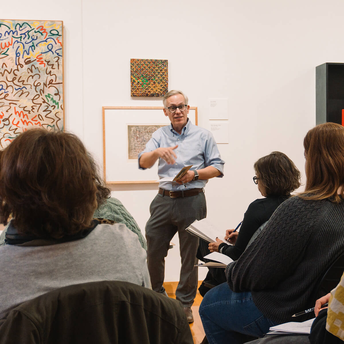 At the front of an art gallery, William Crow lectures to a group of people holding pencils and notepads on their laps. 