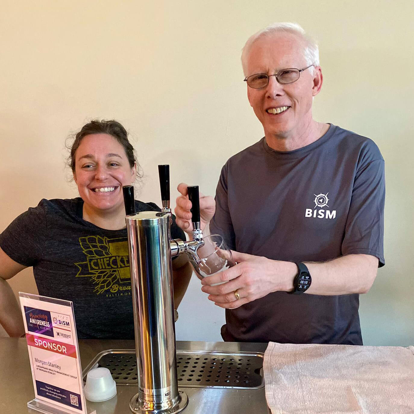 Standing beside a woman behind a beer tap, Mike Gosse pours beer into a pint glass, wearing a dark gray B.I.S.M. t-shirt.