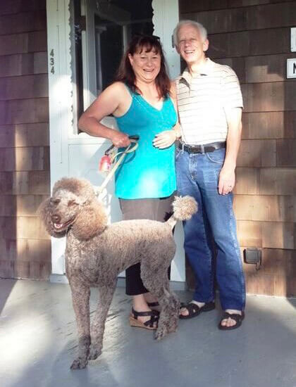 Husband and wife stand side by side on a porch holding their standard poodle on a leash.