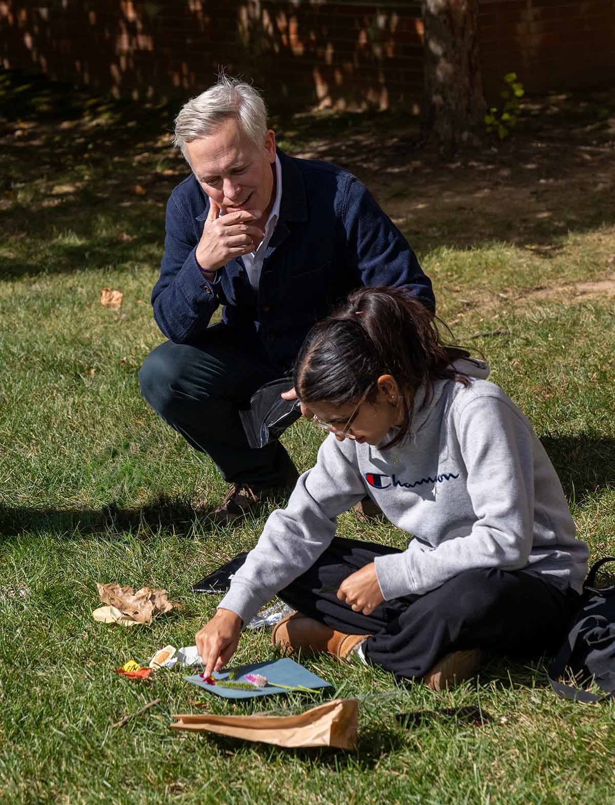 On a sunny autumn day, William Crow squats next to a student sitting in the grass manipulating organic elements onto a piece of blue paper.