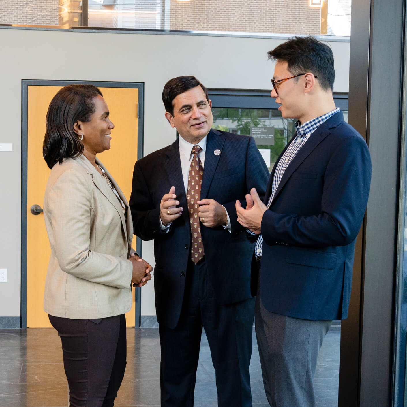 Dean Malhotra stands between a man and woman, all dressed in business attire inside the business building, gesturing with their hands as they talk.