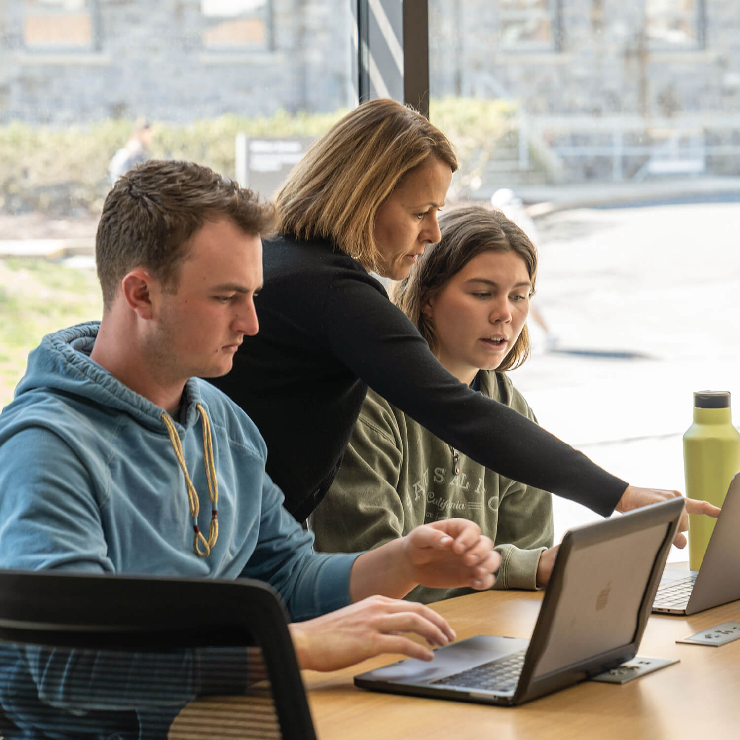 Professor Koukova stands between two students working at a desk on their laptops and leans over one to point at her computer.