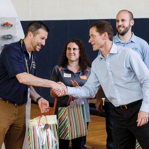 Two men happily lean in to shake hands in front of two colleagues, holding colorful striped gift bags.